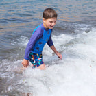 A young boy in a blue star-patterned rash vest and UV Protective Swim Shorts Multi Check, playing happily in the sea on a sunny day.